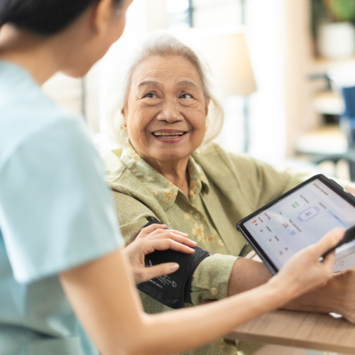 A caregiver checking an older adult’s blood pressure with a digital tablet nearby, while the older adult smiles warmly during an in-home health assessment.