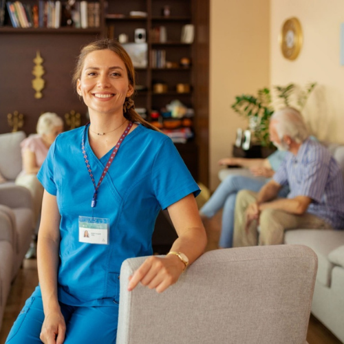 A smiling care worker in blue scrubs standing in a comfortable living room, with older adults socialising in the background, representing professional and friendly home care support.