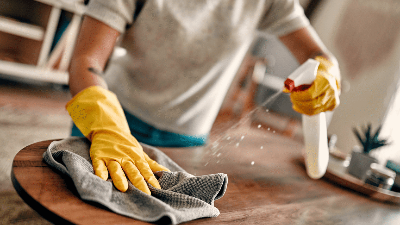 Cleaner wiping and disinfecting a wooden table using gloves and cleaning spray.